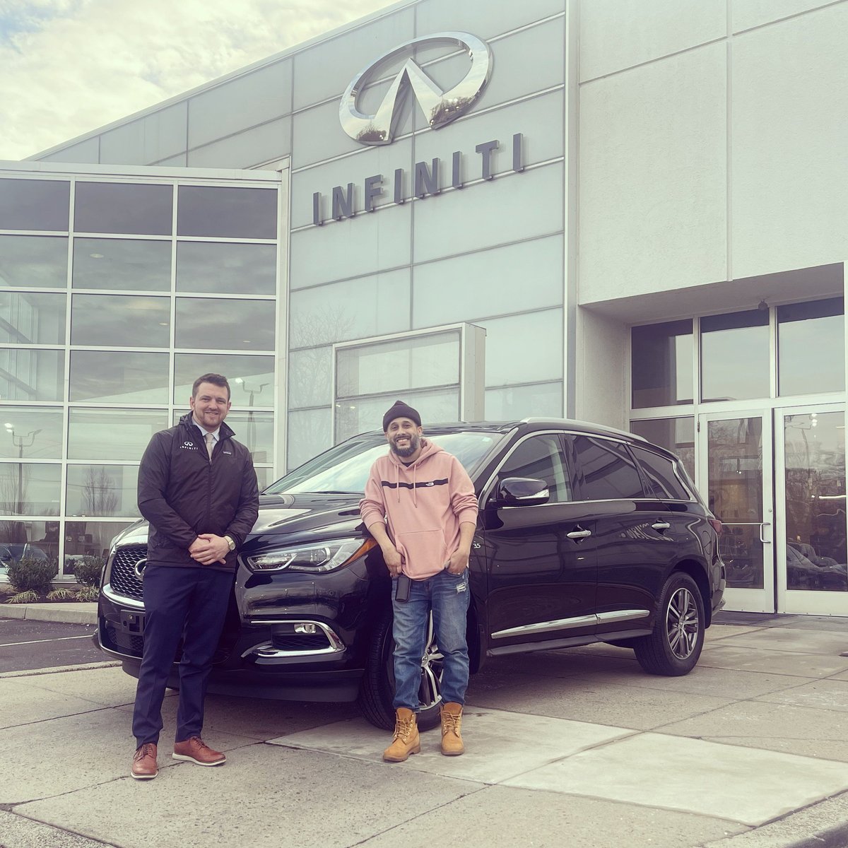 Two men standing in front of a dark SUV outside the INFINITI dealership building