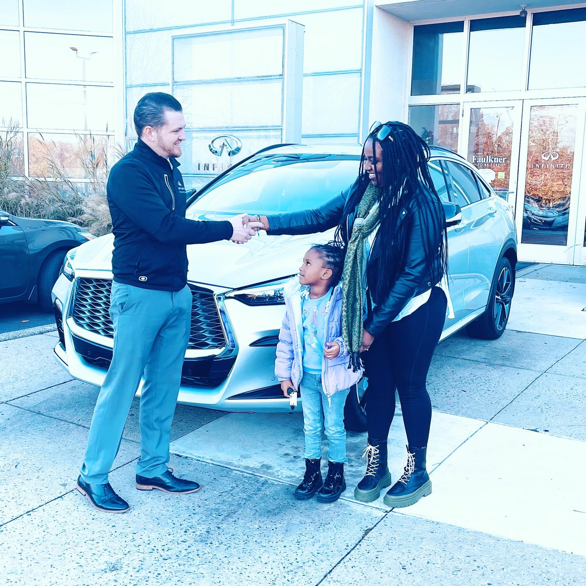 Salesman shaking hands with a mother and young child in front of a blue SUV at the car dealership