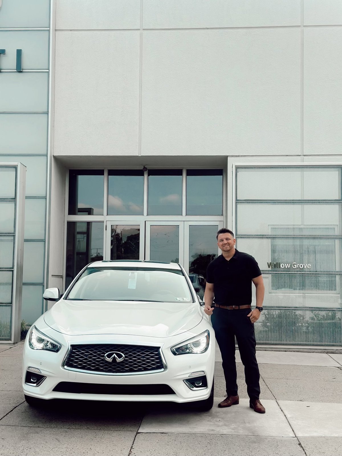Nick Arnott in a black shirt standing next to a white INFINITI QX60 outside the Faulkner INFINITI Willow Grove dealership, with the Willow Grove storefront signage behind him