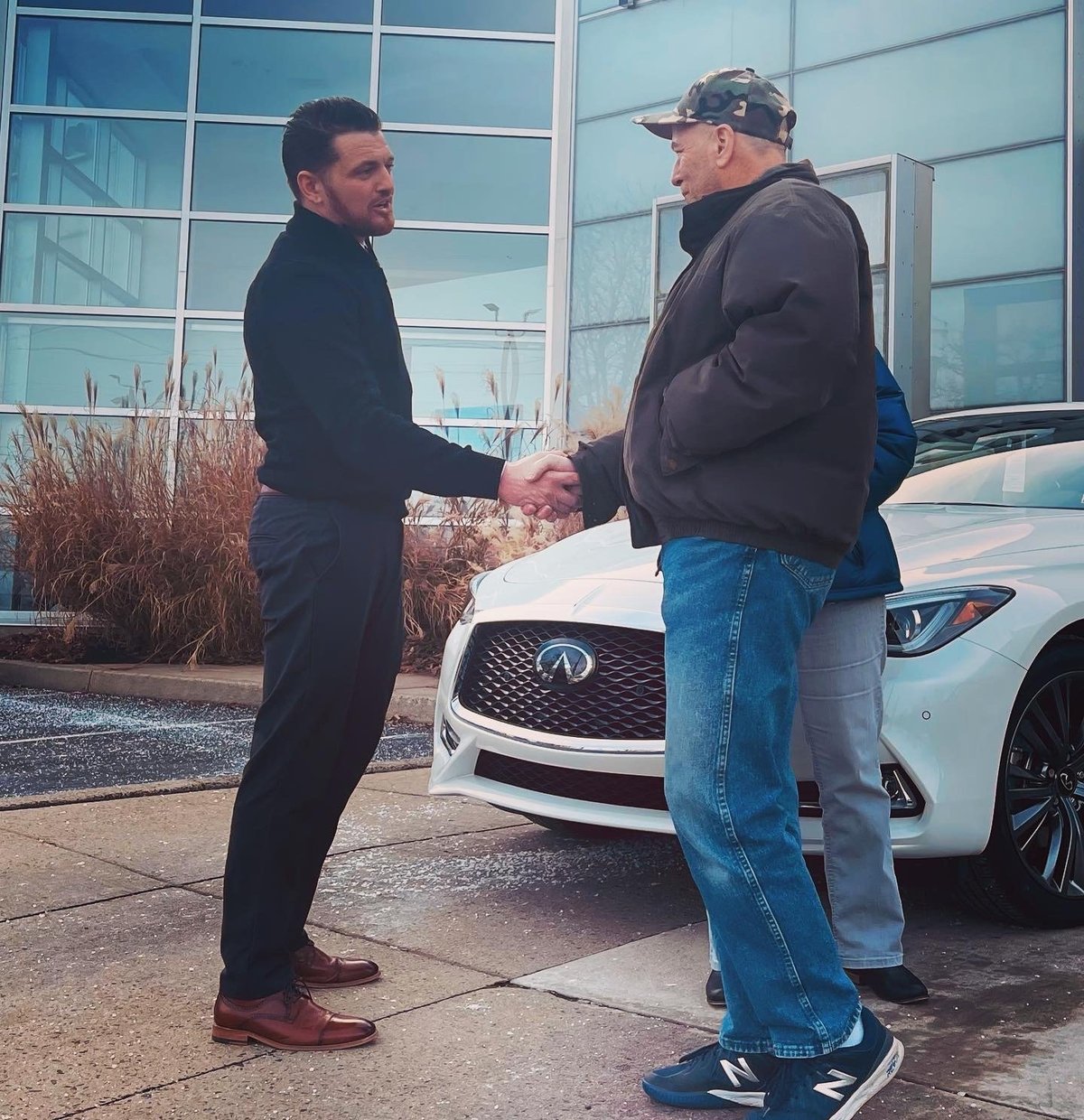 Two men shaking hands in front of a white luxury car at a dealership with modern glass building in background