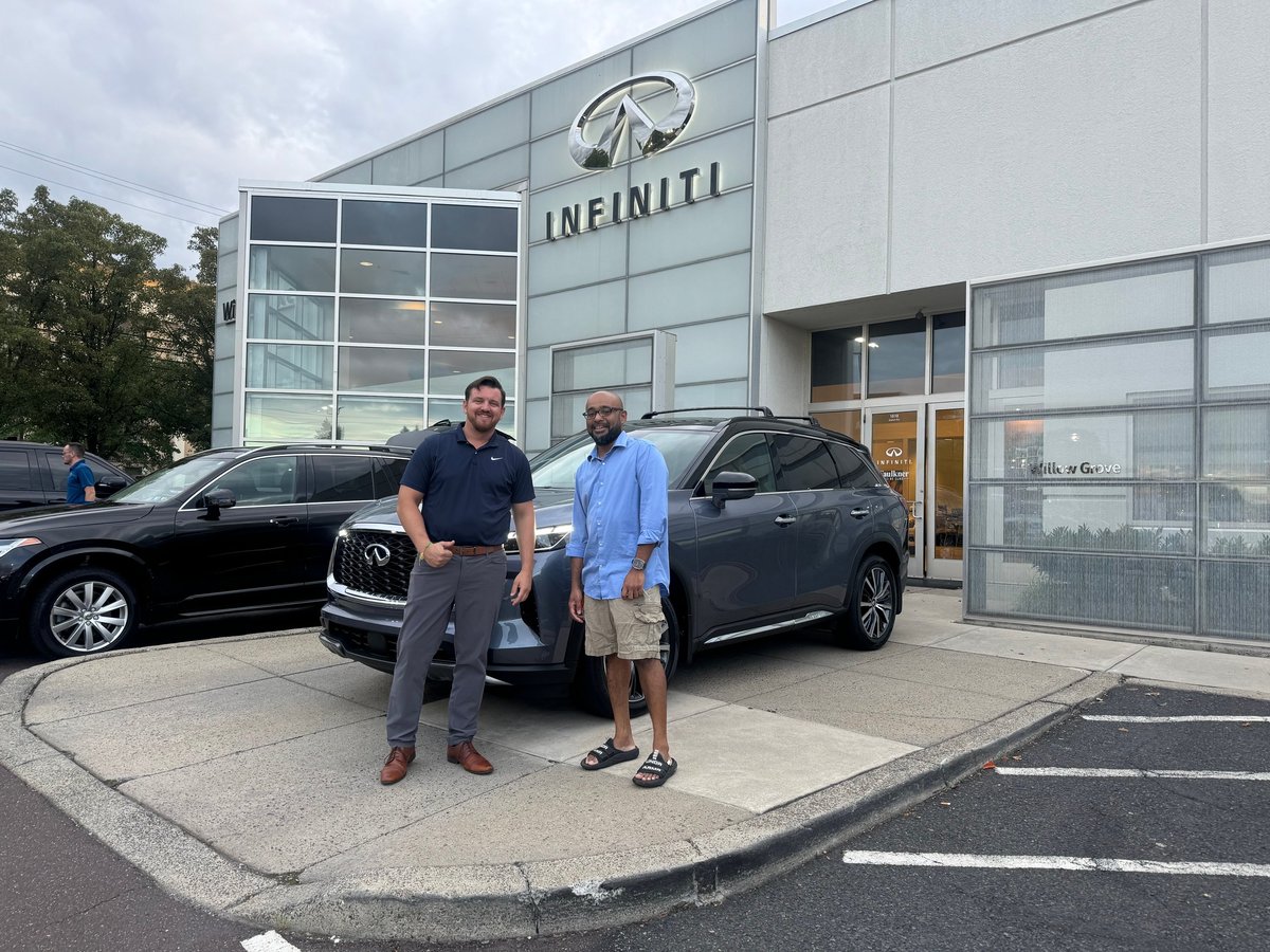 Two men standing between two cars outside the INFINITI dealership with glass front and signage, on delivery day