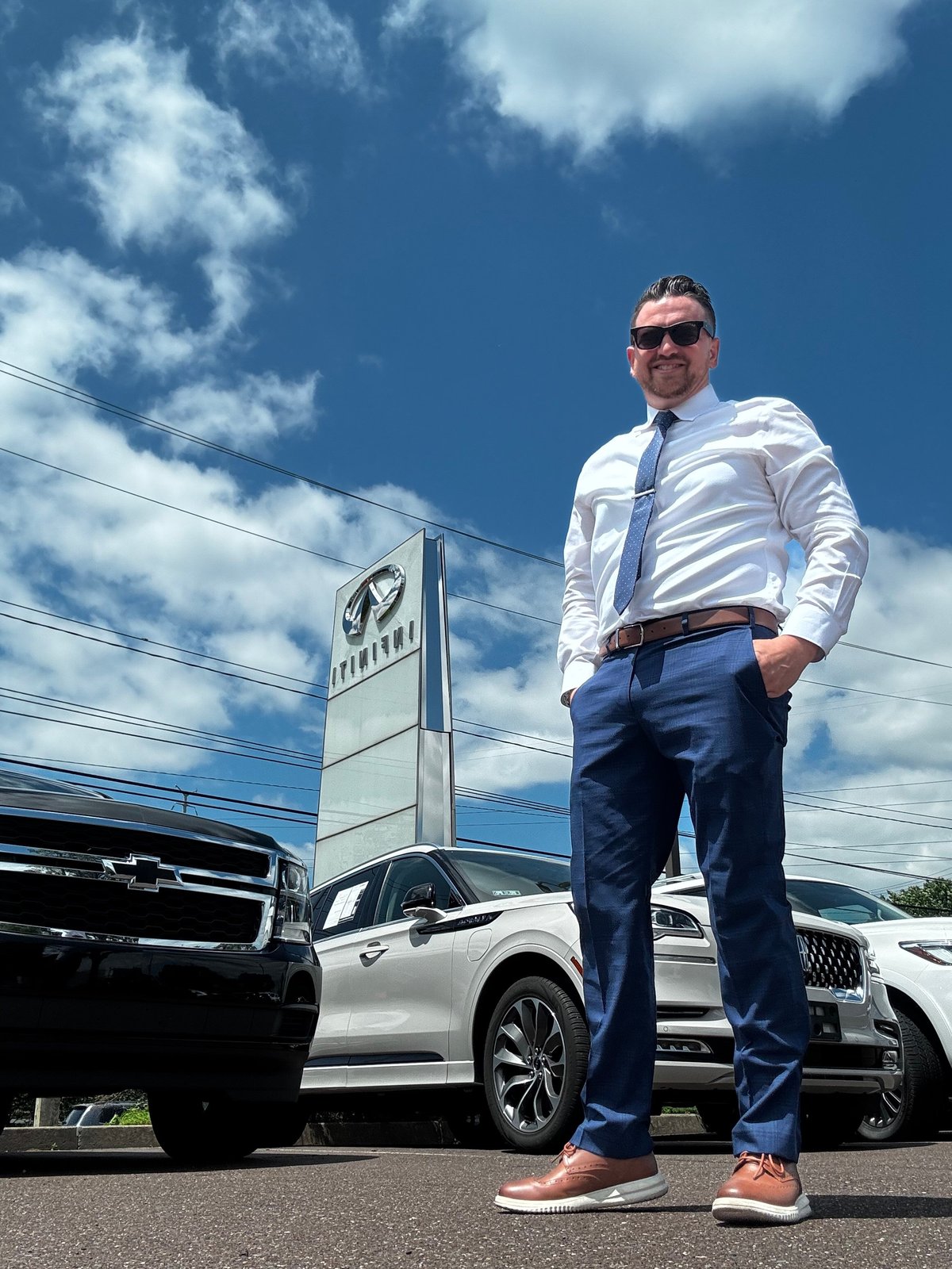 Nick Arnott in business casual attire standing confidently at the INFINITI dealership with vehicles and signage in the background under a blue sky