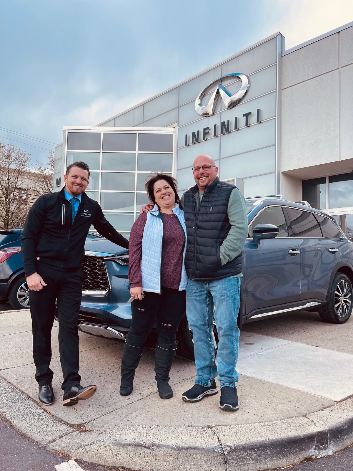 Three people posing in front of an Infiniti dealership with cars parked behind them