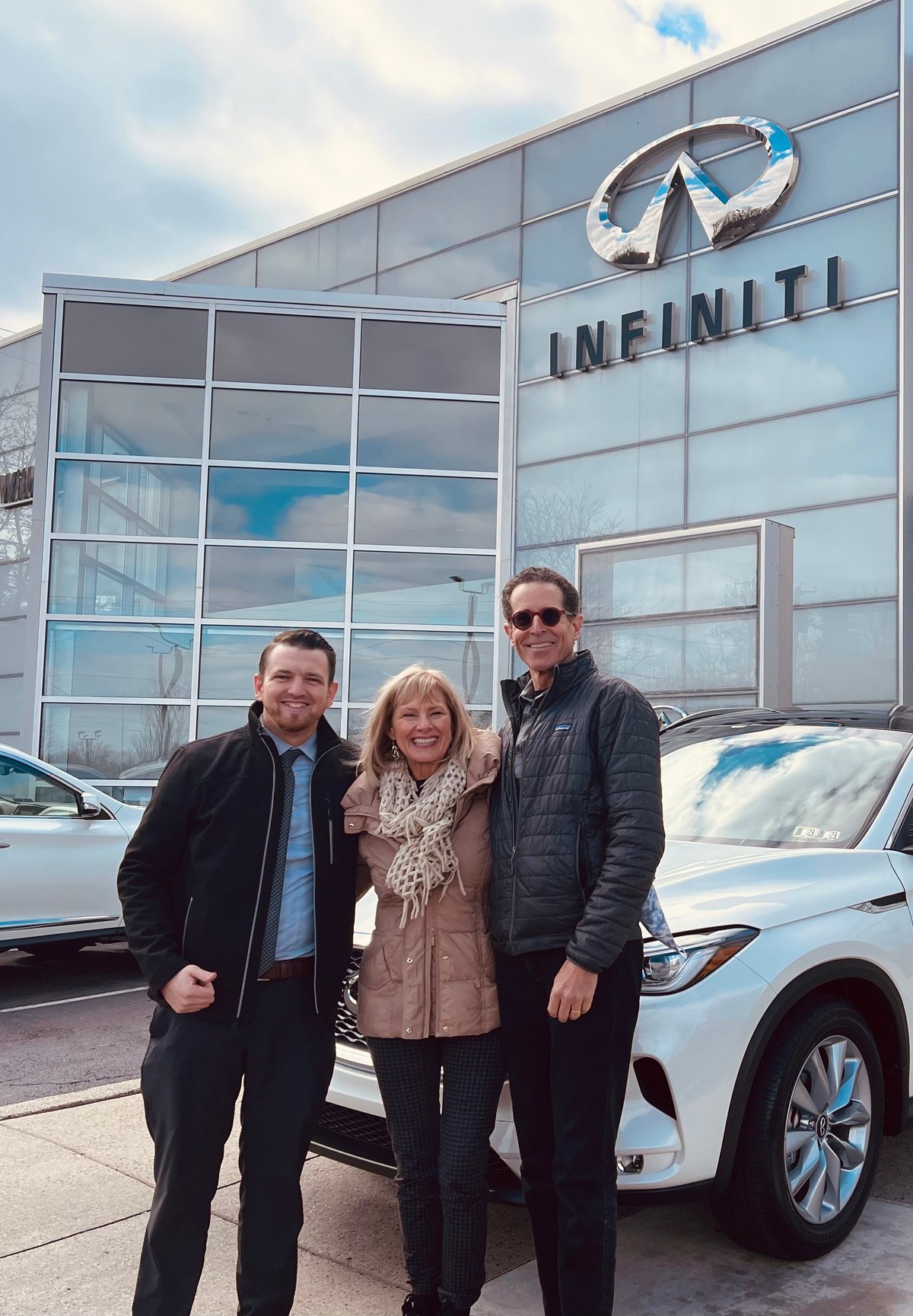 Three people standing in front of an Infiniti dealership with a white luxury vehicle visible in the background