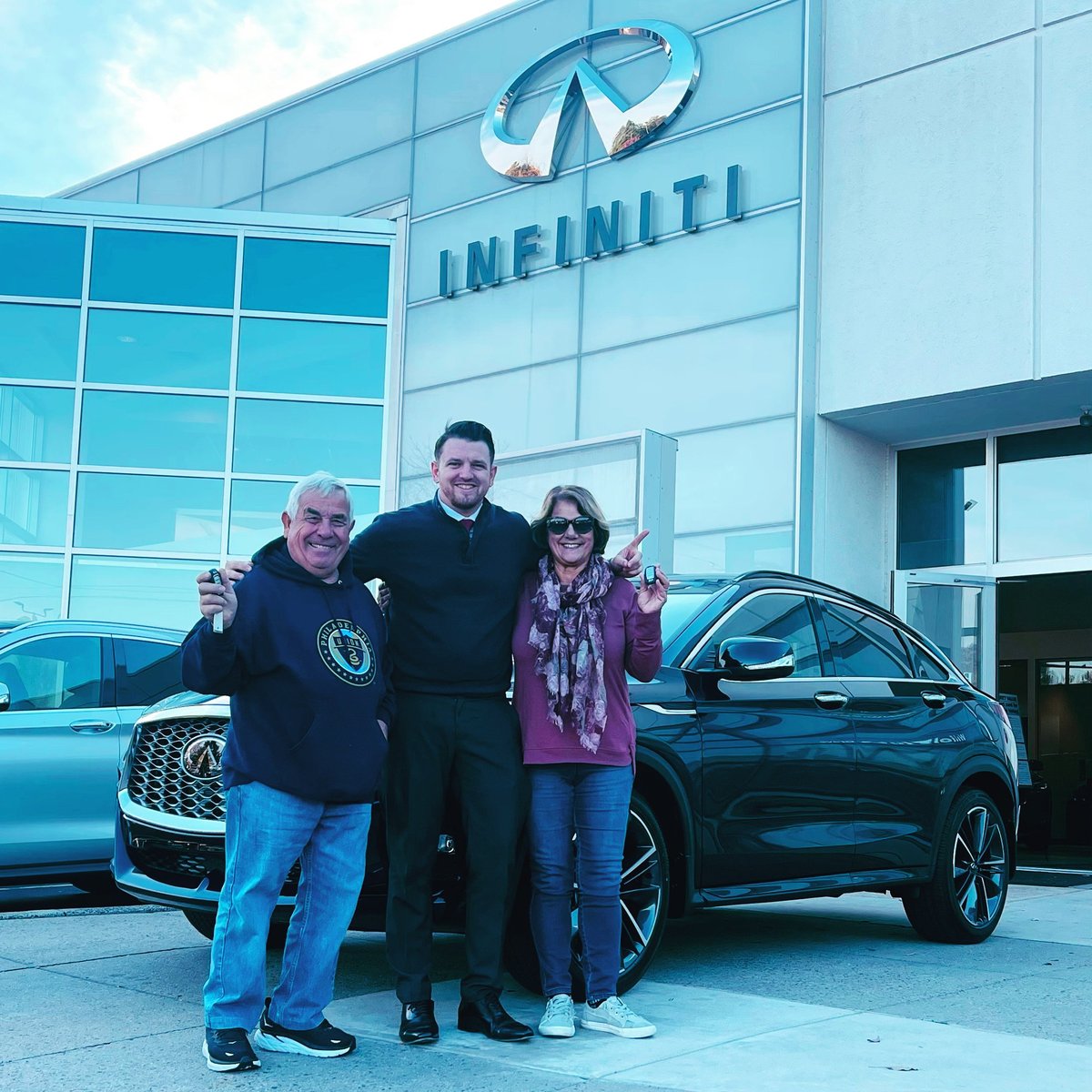 Three people posing in front of the INFINITI dealership with luxury vehicles displayed behind them