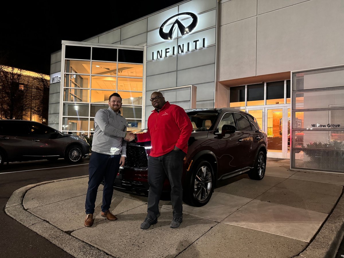 Two men standing beside a dark INFINITI SUV in front of the Faulkner INFINITI Willow Grove dealership at night with illuminated signage